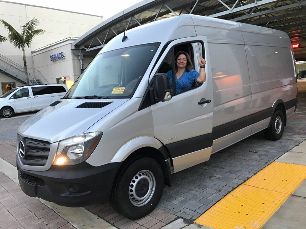 Woman with a blue shirt smiles and gives a thumbs-up from the driver's seat of a silver van.
